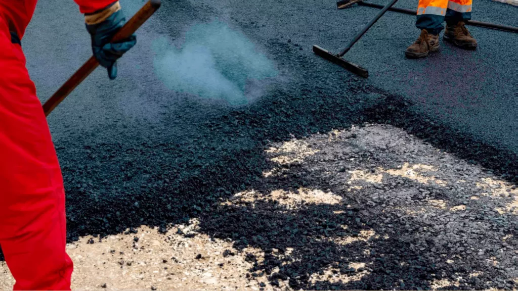 Road workers in high-visibility gear repair a dark asphalt surface with steaming hot tar, using shovels; the scene feels focused and industrious.