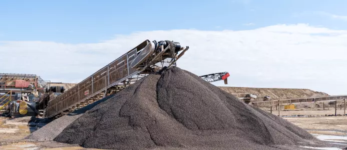 A conveyor system pours dark gravel onto a large aggregate pile at an industrial quarry, beneath a bright blue sky; the scene feels busy and expansive.