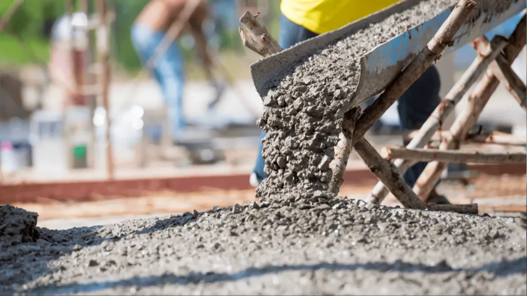 Workers pour wet concrete from a metal chute onto a construction site, forming a rough gray slab; bright daylight conveys active, industrious work.